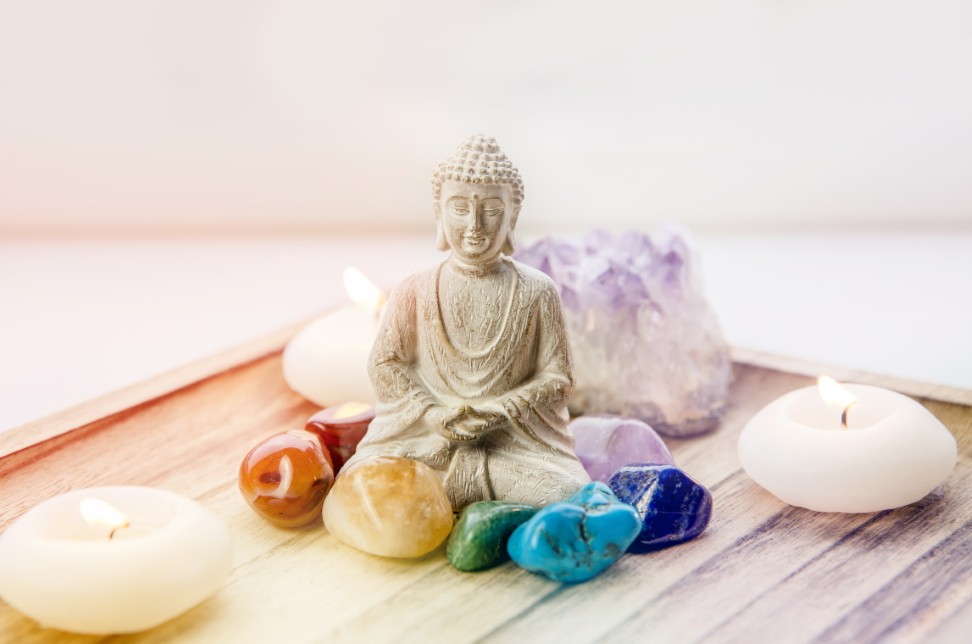 Meditierende Buddha-Statue auf Holzfläche, umgeben von bunten Edelsteinen und brennenden Kerzen, im Hintergrund ein Amethyst-Kristall.